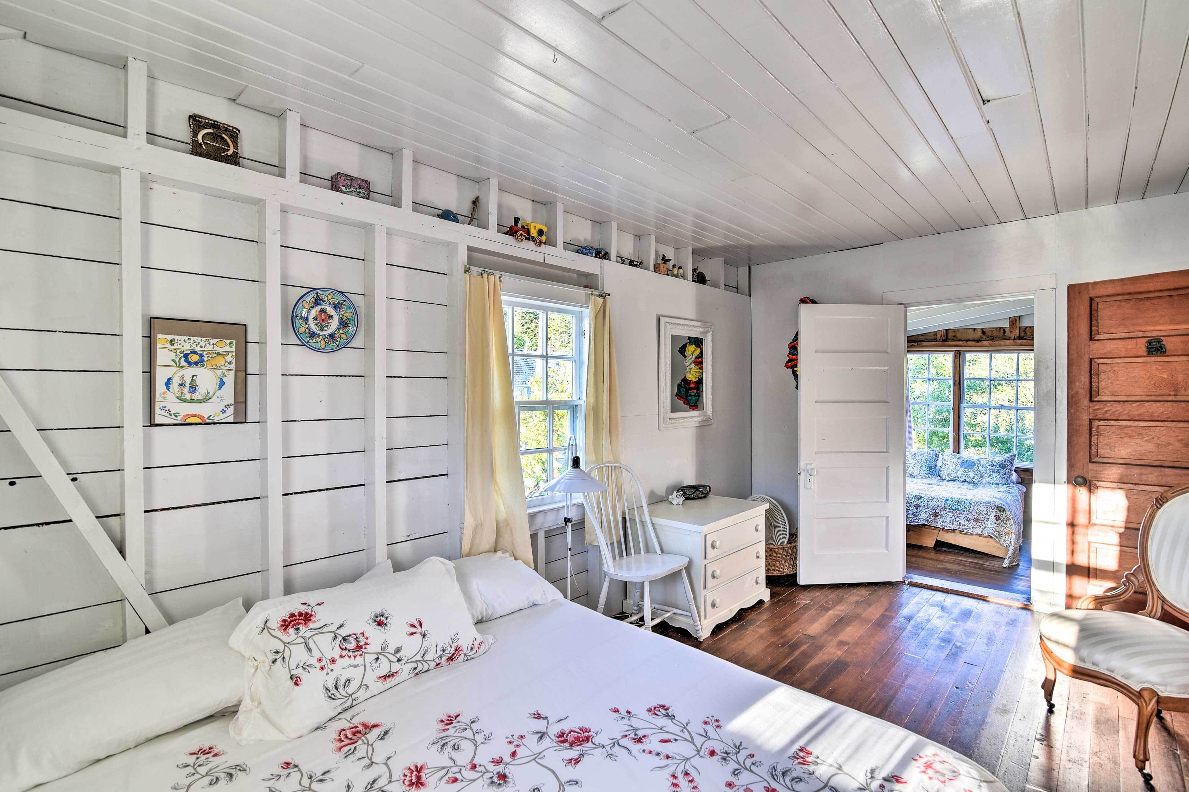 Een slaapkamer met een bed, een bureau en een stoel. Er is een raam met gordijnen, een deur naar een andere kamer, en een houten wand met planken.