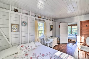 Une chambre à coucher avec un lit, un bureau et une chaise. Il y a une fenêtre avec des rideaux, une porte qui donne sur une autre pièce et un mur en bois avec des étagères.