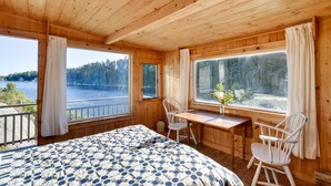 Eine Holzhütte mit Bett, Tisch, zwei Stühlen und großen Fenstern mit Blick auf einen See und Wald.