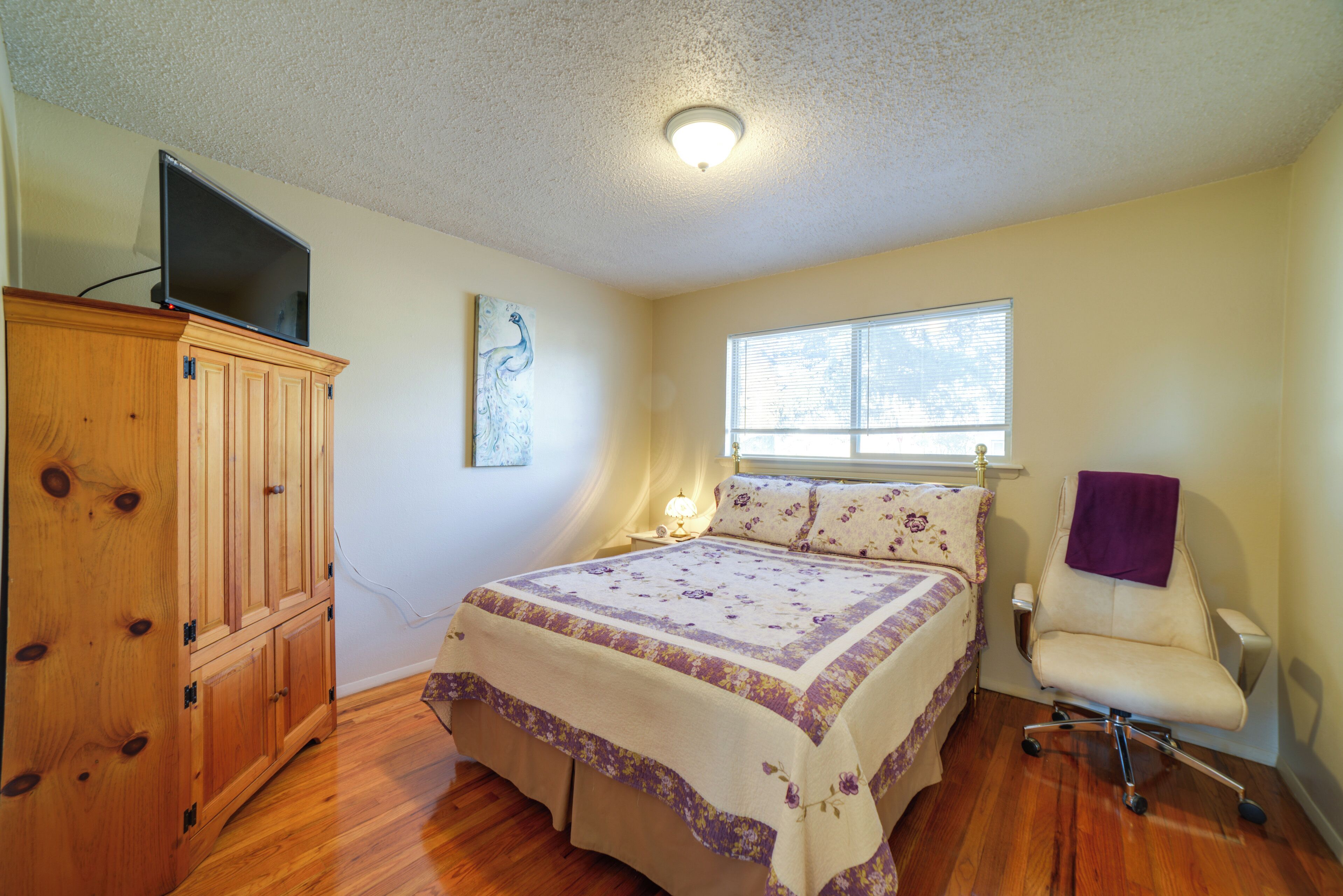 Un dormitorio con un armario de madera, una cama con ropa de cama floral, una silla y una ventana con persianas.
