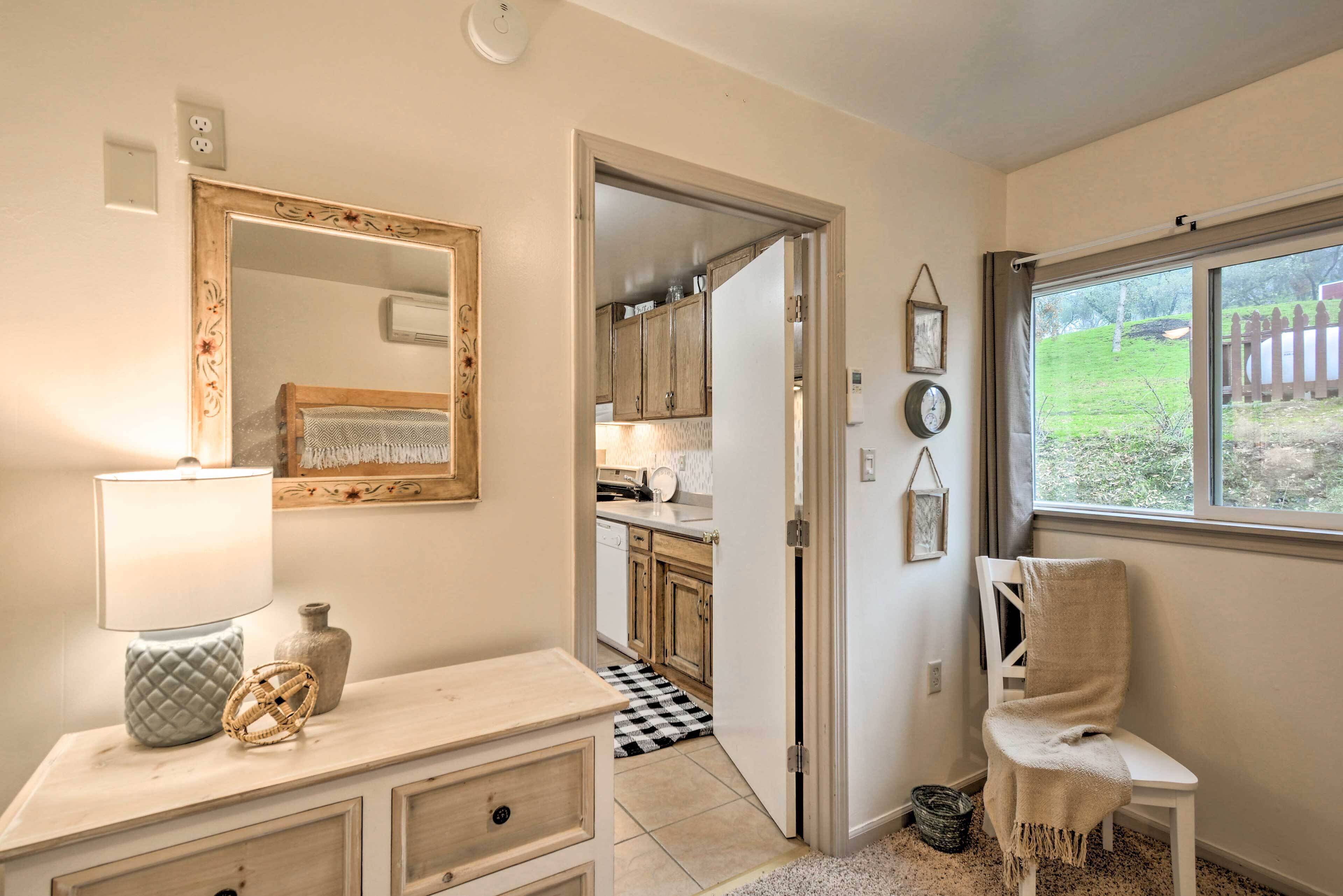 A room with a wooden dresser, a mirror, a lamp, a chair, and a window with a view of a garden.