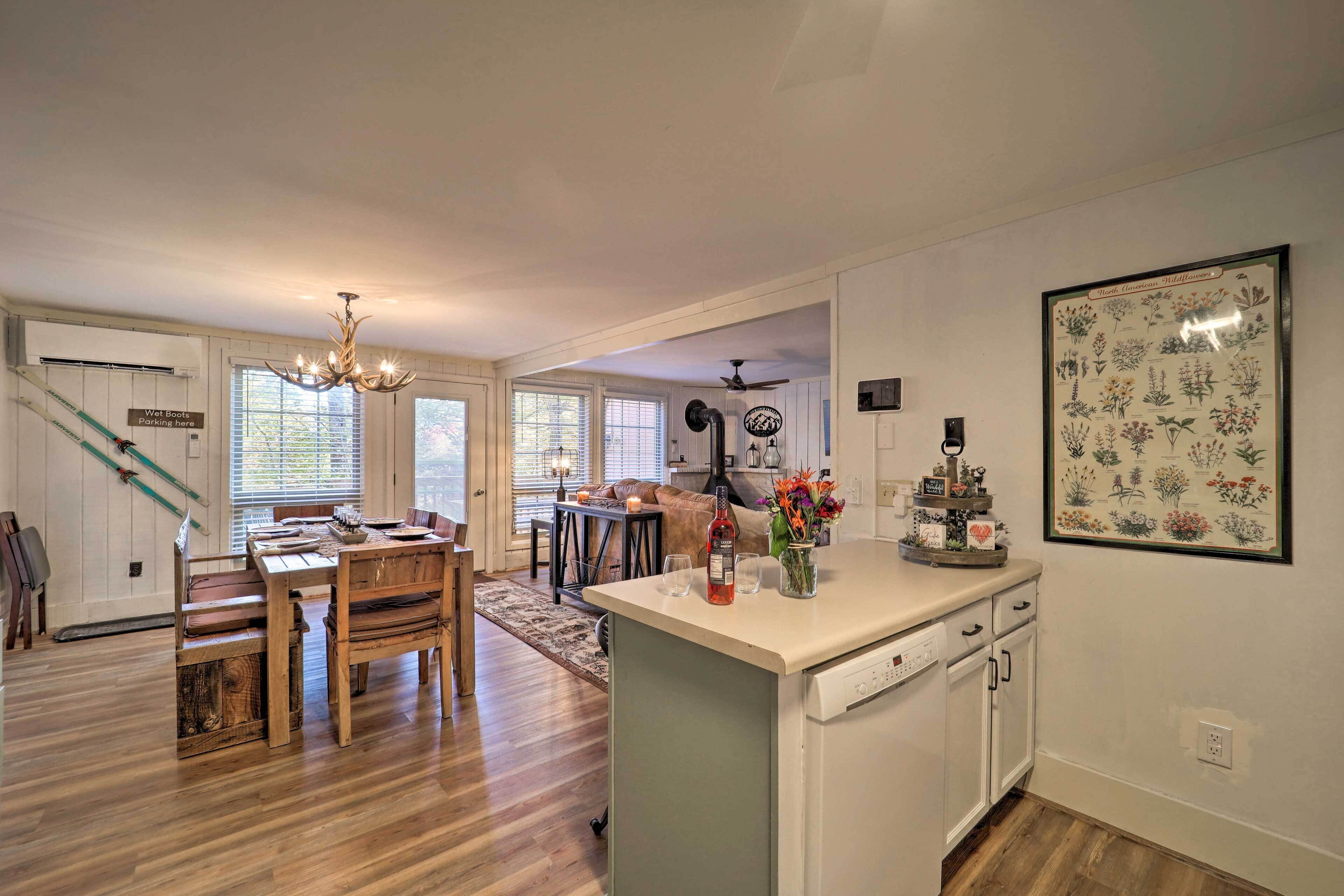 A spacious kitchen with a central island, wooden dining table, and chairs. There is a wet boots parking sign on the wall, a ceiling fan, and a framed botanical illustration.
