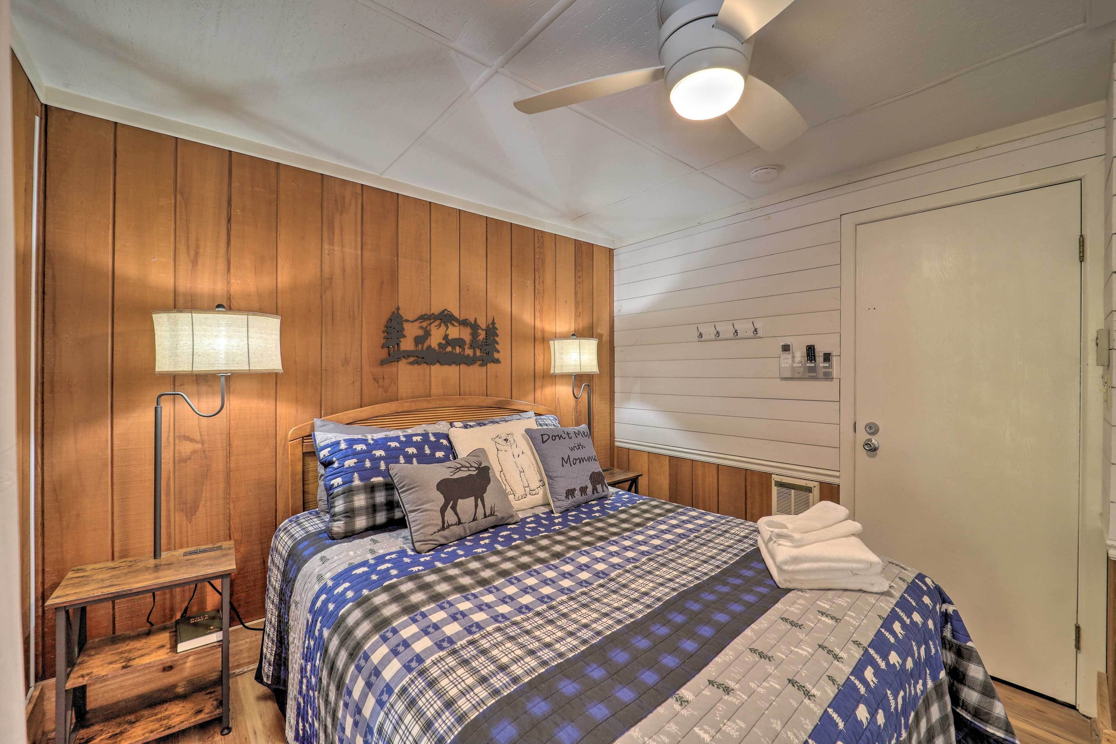 A bedroom with a wooden wall, a bed with a blue and white checkered bedspread, two bedside lamps, a ceiling fan, and a door with a handle.