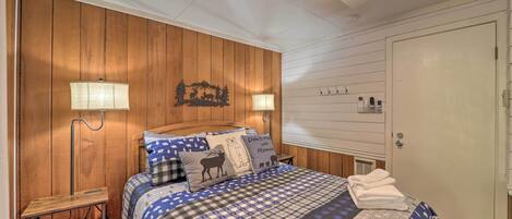 A bedroom with a wooden wall, a bed with a blue and white checkered bedspread, two bedside lamps, a ceiling fan, and a door with a handle.