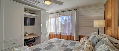 A bedroom with a large bed, a ceiling fan, a TV on a shelf, and a window with curtains.