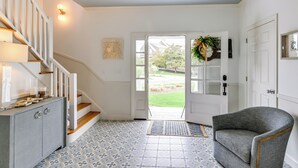 A hallway with a staircase, a door leading outside, a grey armchair, and patterned floor tiles. - Stunning Westhampton Beach Home w/ Private Pool (Westhampton Beach)