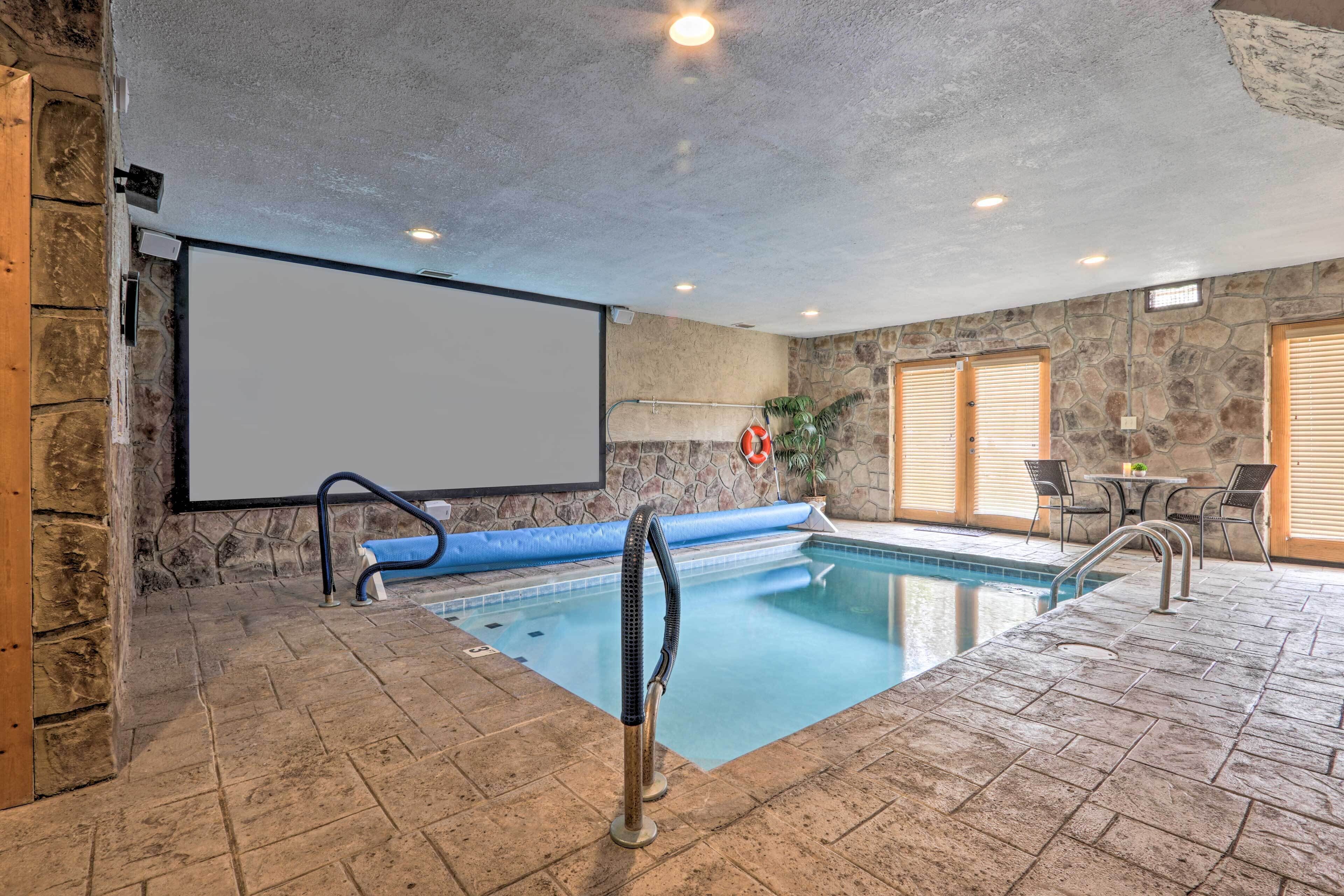 An indoor swimming pool with a stone wall, a projector screen, and a seating area with chairs and a table.