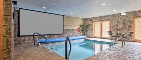An indoor swimming pool with a stone wall, a projector screen, and a seating area with chairs and a table.