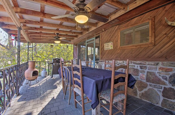 A covered outdoor patio with wooden tables and chairs, a stone wall, and a rustic ceiling fan.