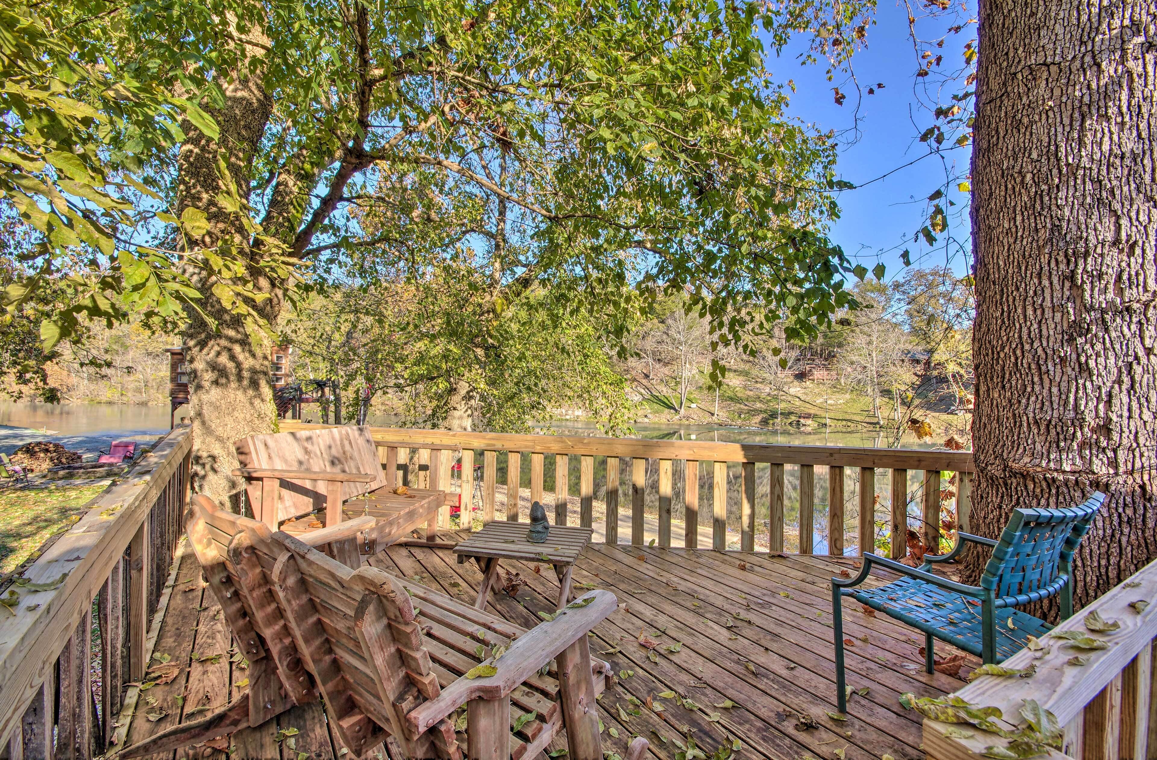 Une terrasse en bois avec des bancs et une table, donnant sur une étendue d’eau et entourée d’arbres.