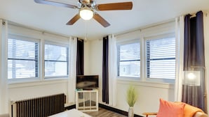 Una sala de estar moderna con ventilador de techo, televisor de pantalla plana sobre un mueble bajo, una mesa de centro blanca, un sofá con un cojín naranja y ventanas amplias con persianas.