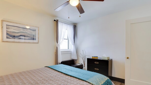 Un dormitorio con cama, ventilador de techo, ventana con cortinas, radiador, cómoda y puerta.