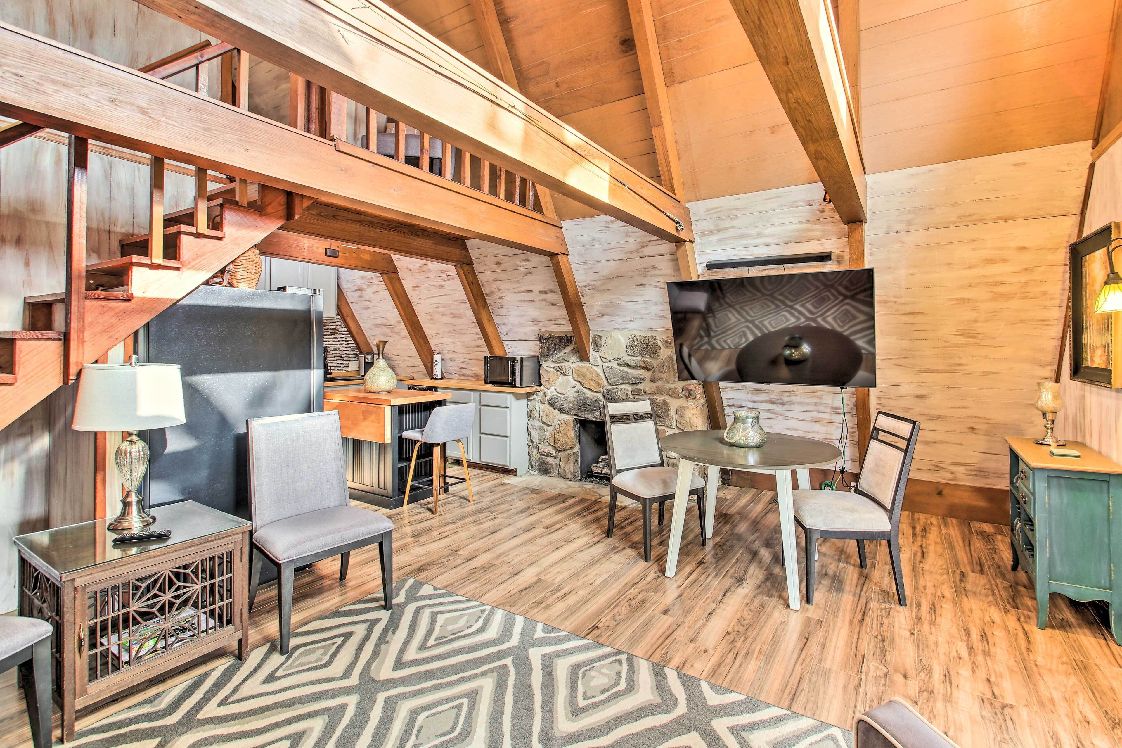 A spacious loft-style room with a high ceiling, exposed wooden beams, a stone fireplace, and a dining area with modern furniture.