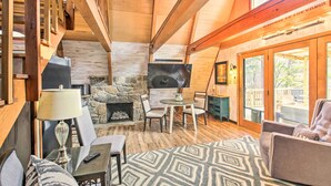 A cozy living room with a stone fireplace, wooden beams, and a large window overlooking a deck. - Lake Hamilton Cabin: Boating Getaway! (Hot Springs)