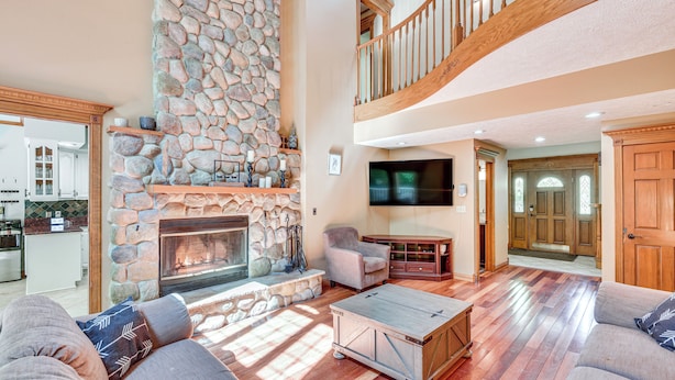 A spacious living room with a stone fireplace, wooden flooring, and a high ceiling with a wooden staircase. - Huge Family Home w/ Pool - 13 Mi to Lake Michigan! (La Porte)