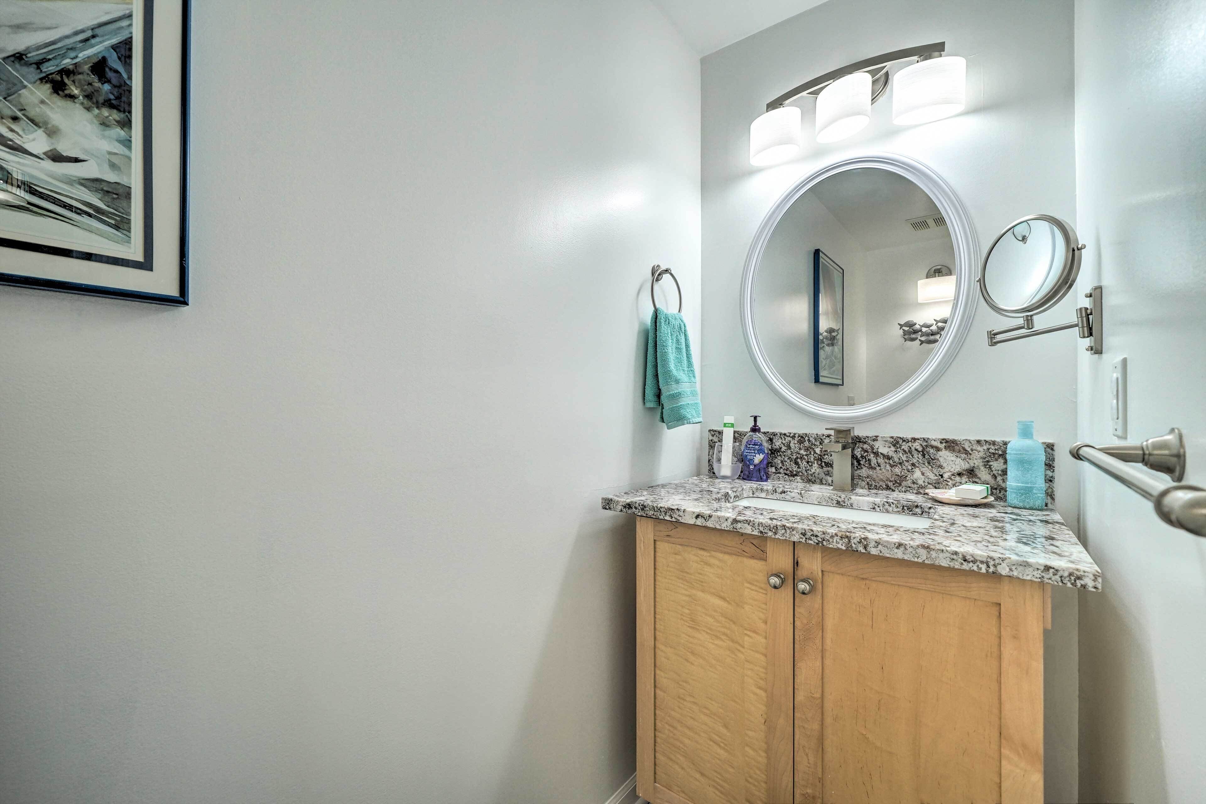 A bathroom with a granite countertop, a wooden cabinet, a round mirror, and a framed artwork on the wall.