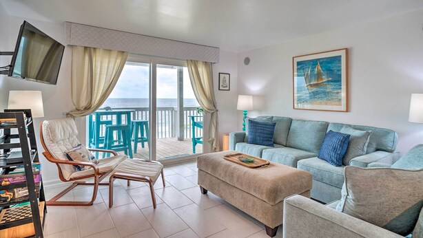 Una sala de estar con sofá, reposapiés y sillón, un televisor, y vista al mar a través de amplias ventanas.