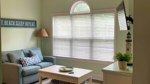 A living room with a light blue sofa, a white coffee table, and a large window with blinds.