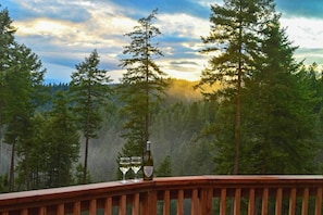 A wooden deck with a bottle of wine and two glasses, overlooking a forested landscape with tall trees and a cloudy sky. - Unique 40 Acre Nature Retreat w/ Trails & Views! (Coeur d'Alene)