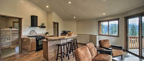 A spacious living area with a kitchen, wooden flooring, and large windows offering a view of the outdoors.