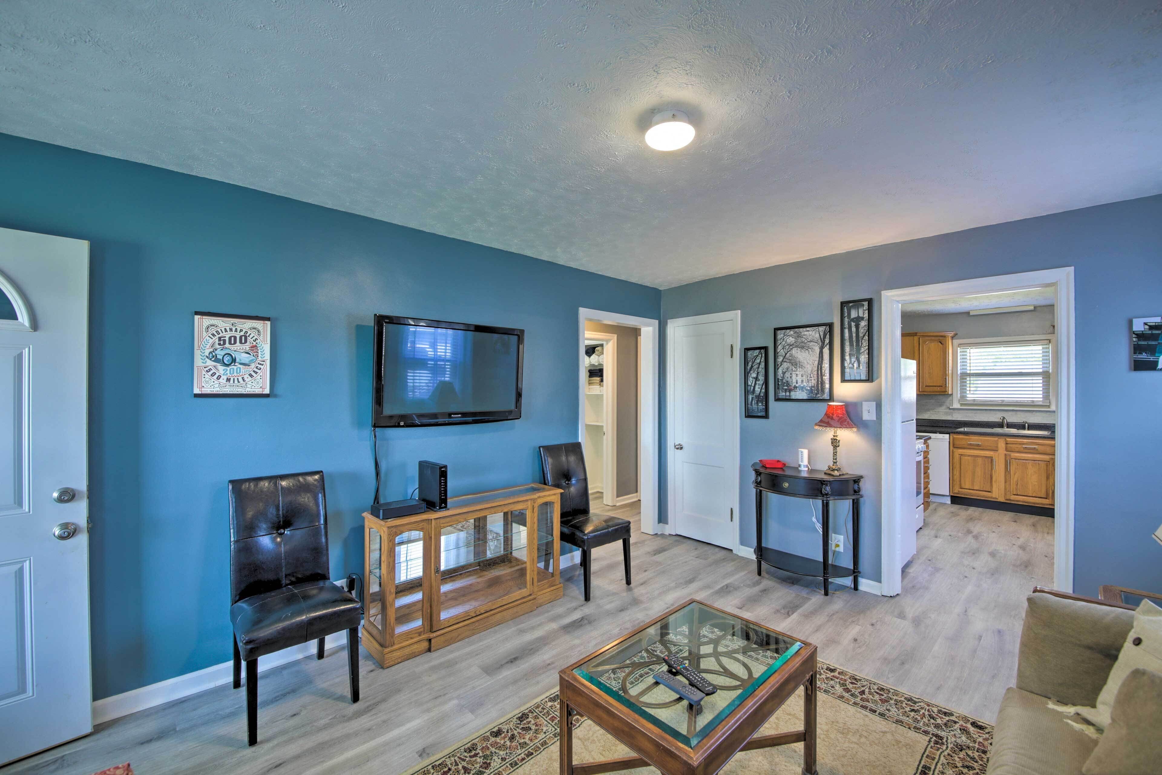 A living room with a blue wall, a television, a glass coffee table, and a kitchen in the background.