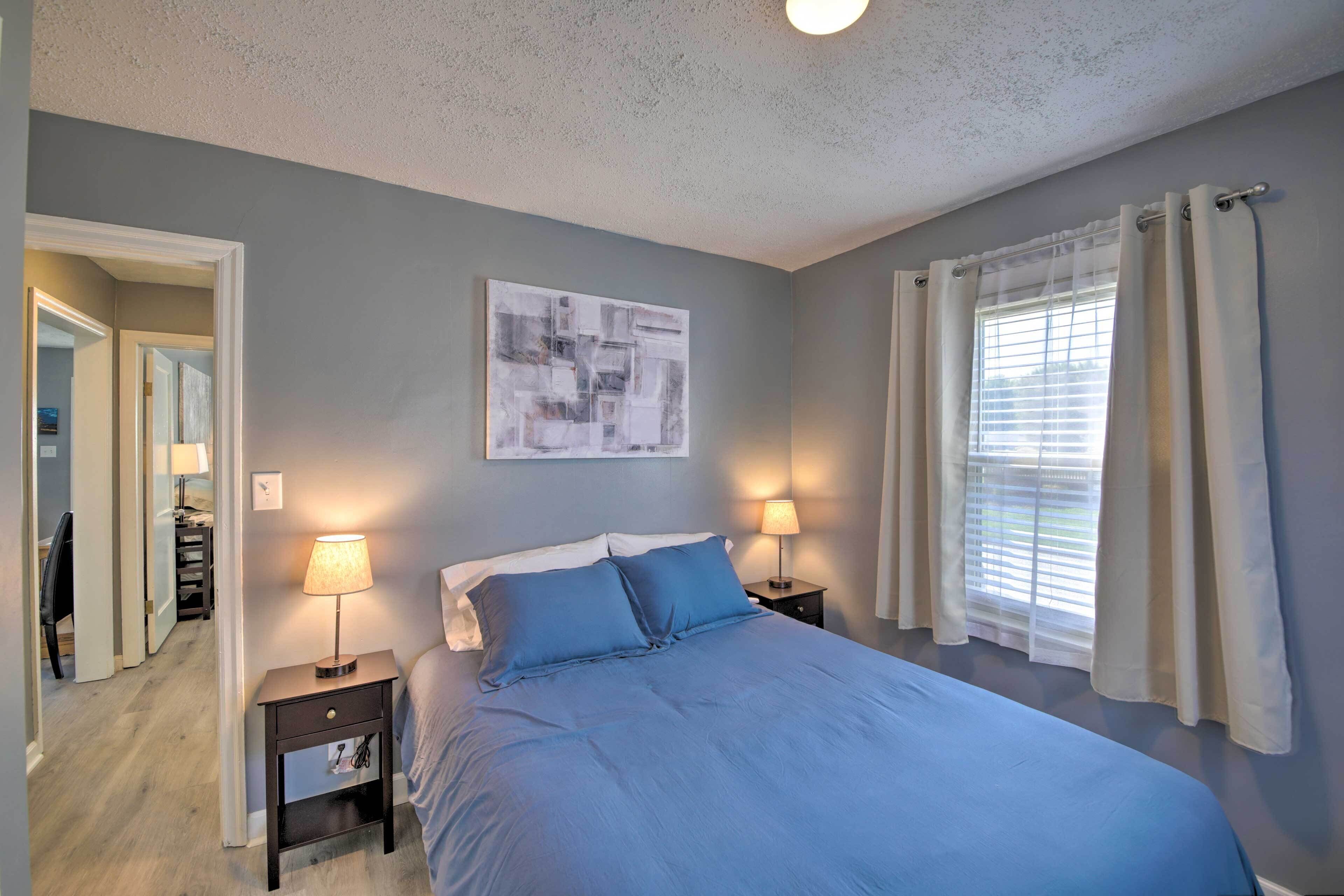A bedroom with a bed, two bedside tables with lamps, a window with curtains, and a wall-mounted abstract art piece.