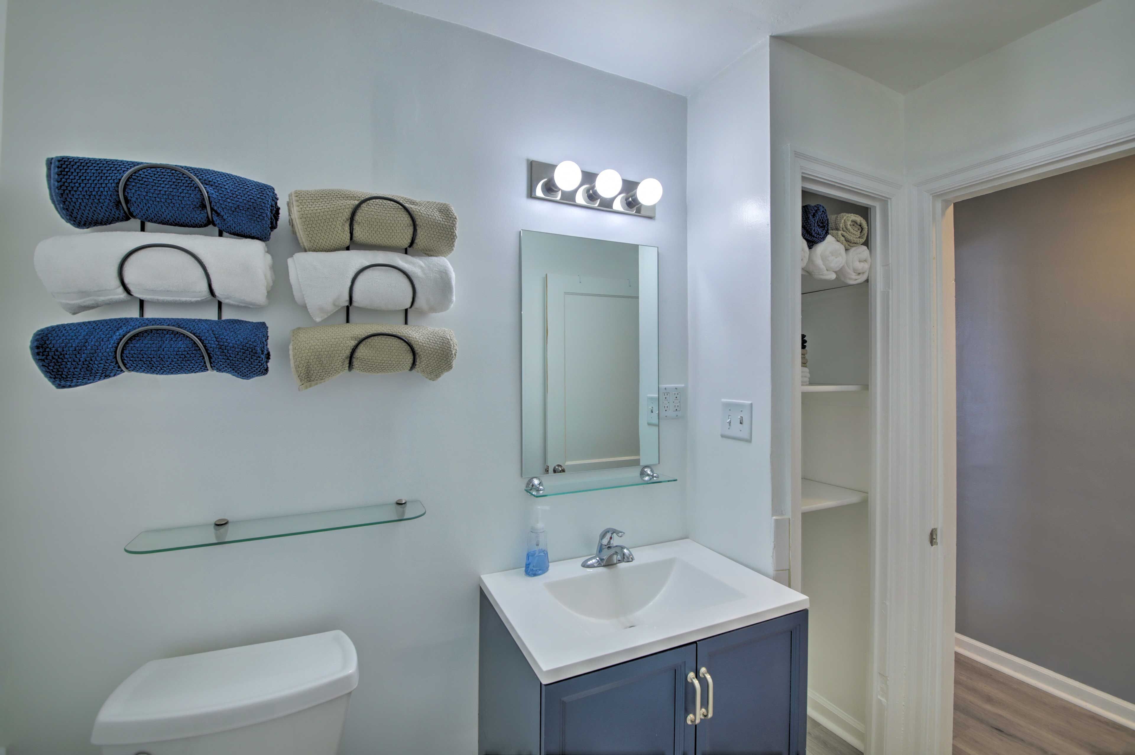 A bathroom with a white toilet, a sink with a glass shelf, a mirror with lights, and towels on a rack.