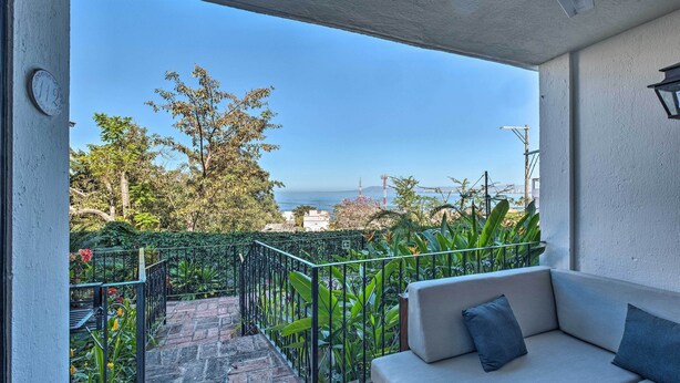 A balcony with a white sofa, a clock, and a view of the sea and trees. - Sweet & Chic Puerto Vallarta Condo: Walk to Beach! (Puerto Vallarta)