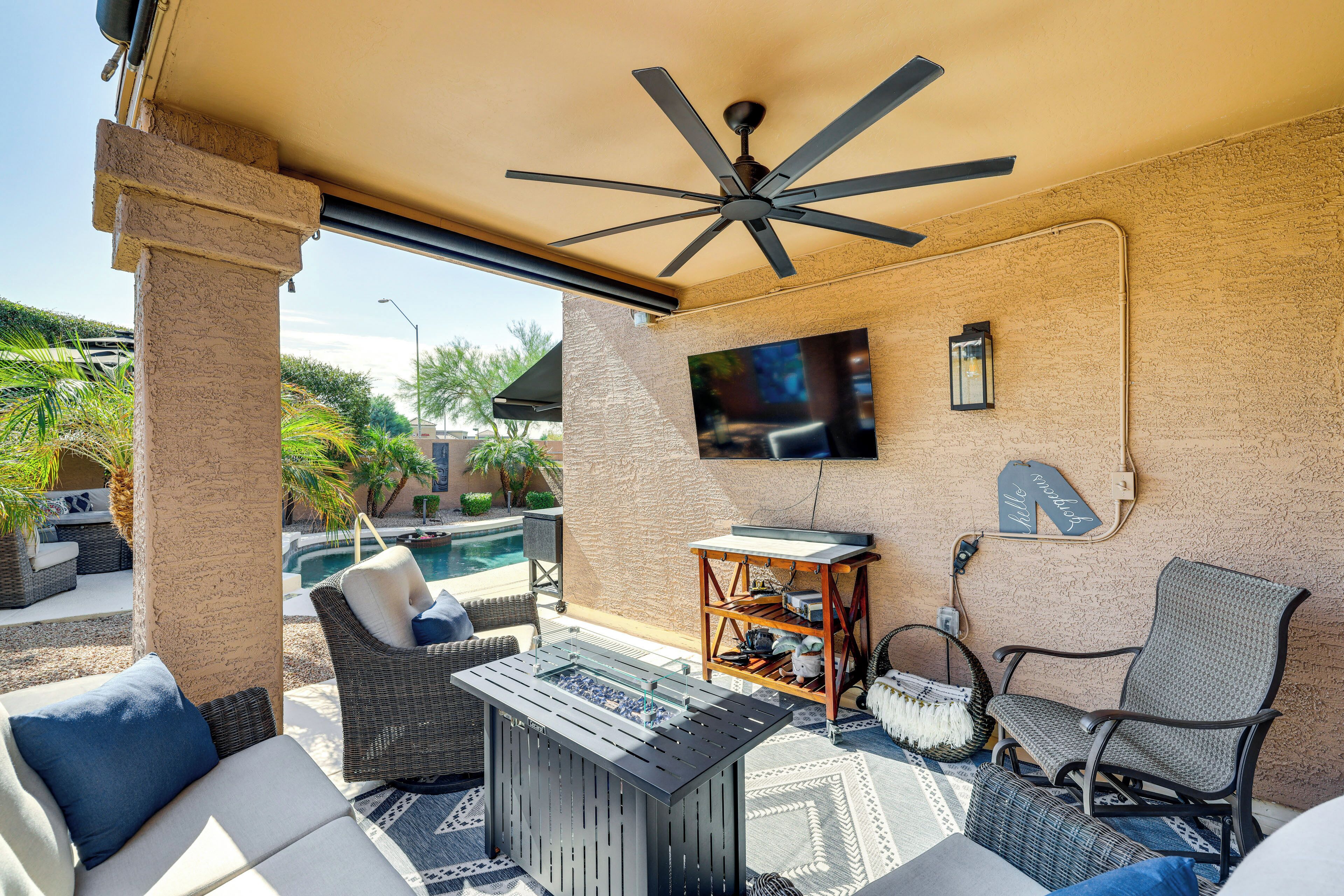 Una veranda esterna riparata con ventola a soffitto, una TV a schermo piatto montata a parete, una piscina e mobili da esterno tra cui un divano, una poltrona e un tavolo.