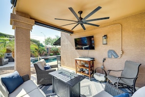 A covered outdoor patio with a ceiling fan, a flat-screen TV mounted on the wall, a pool, and outdoor furniture including a sofa, a chair, and a table.