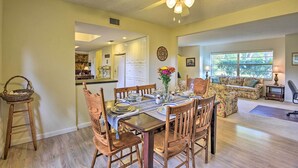 A dining area with a wooden table set for six, surrounded by matching chairs. A ceiling fan is above the table, and a window provides natural light.