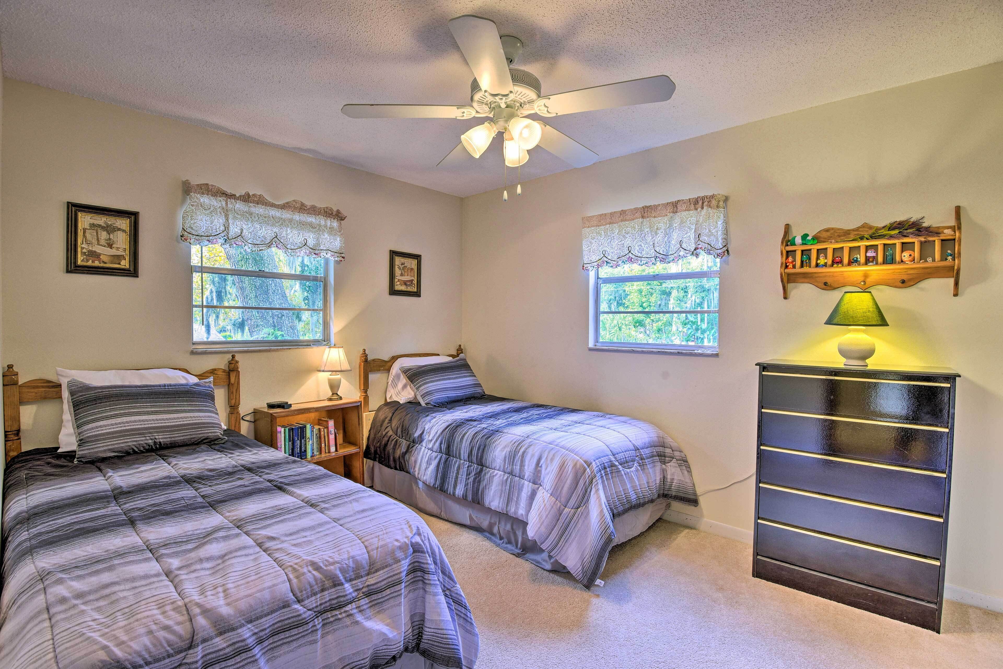 A bedroom with two beds, a ceiling fan, a dresser, and a window with lace curtains.