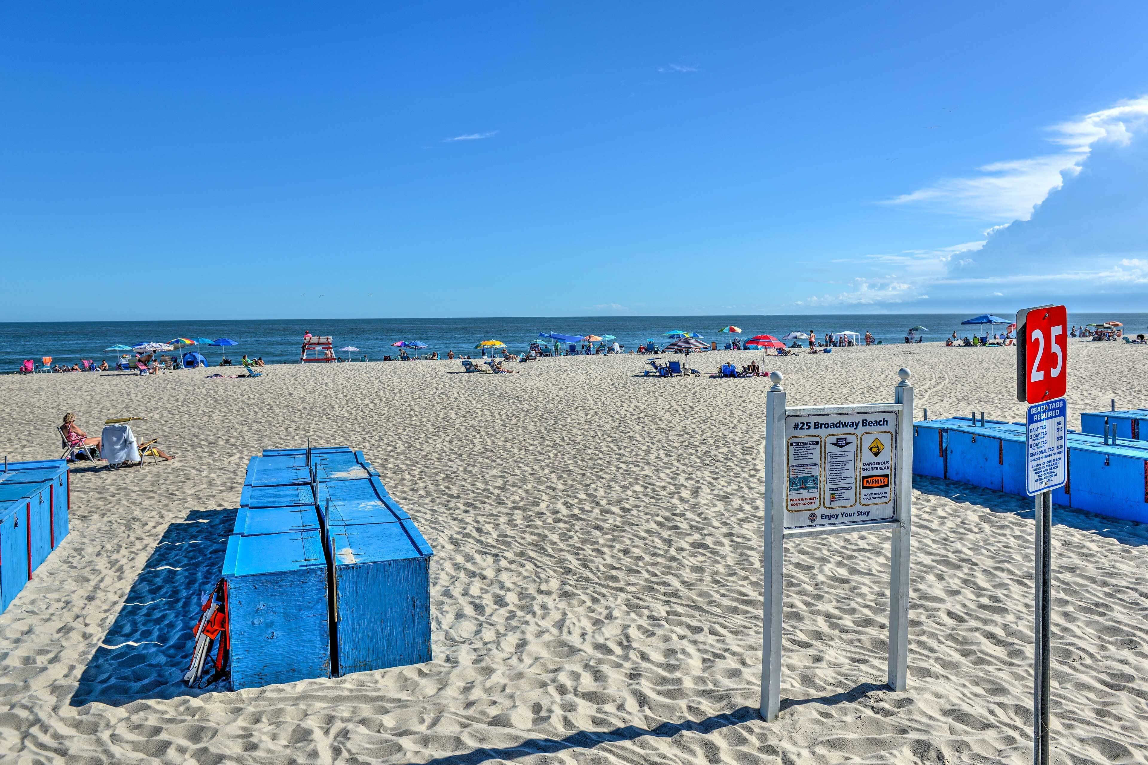 Ein Strand mit blauen Strandhütten, einem Schild mit der Aufschrift '25 Broadway Beach' und Menschen, die unter Sonnenschirmen entspannen.