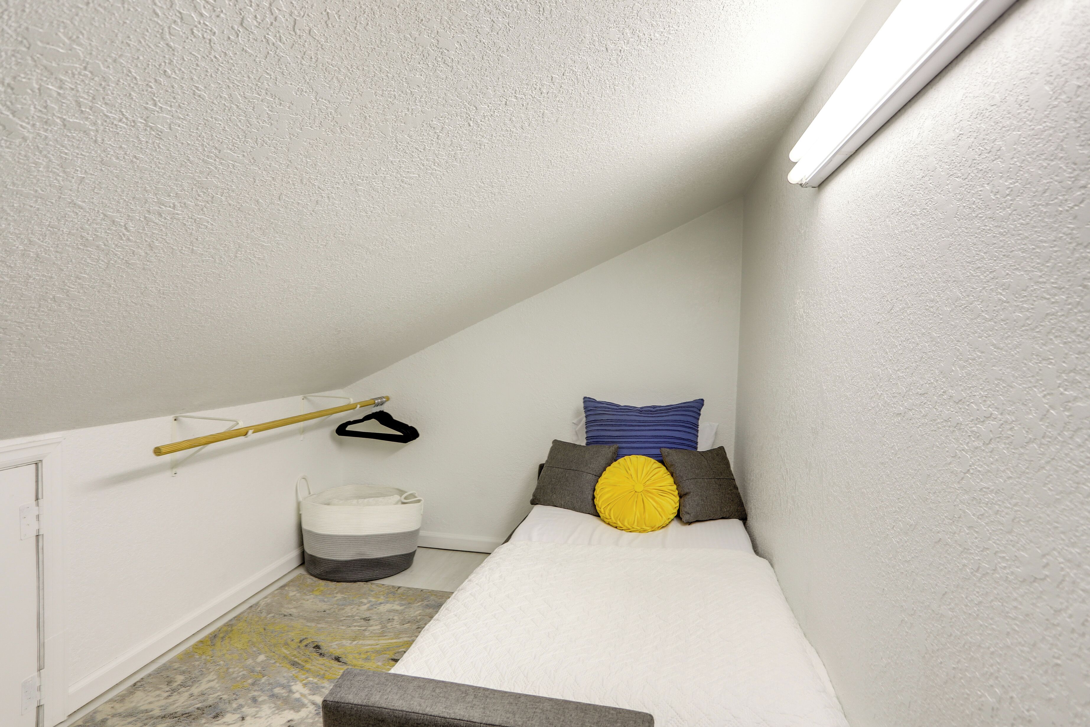 Una habitación pequeña y estrecha con techo inclinado, una cama con colchón y almohadas blancas, una mesita y una lámpara colgante.