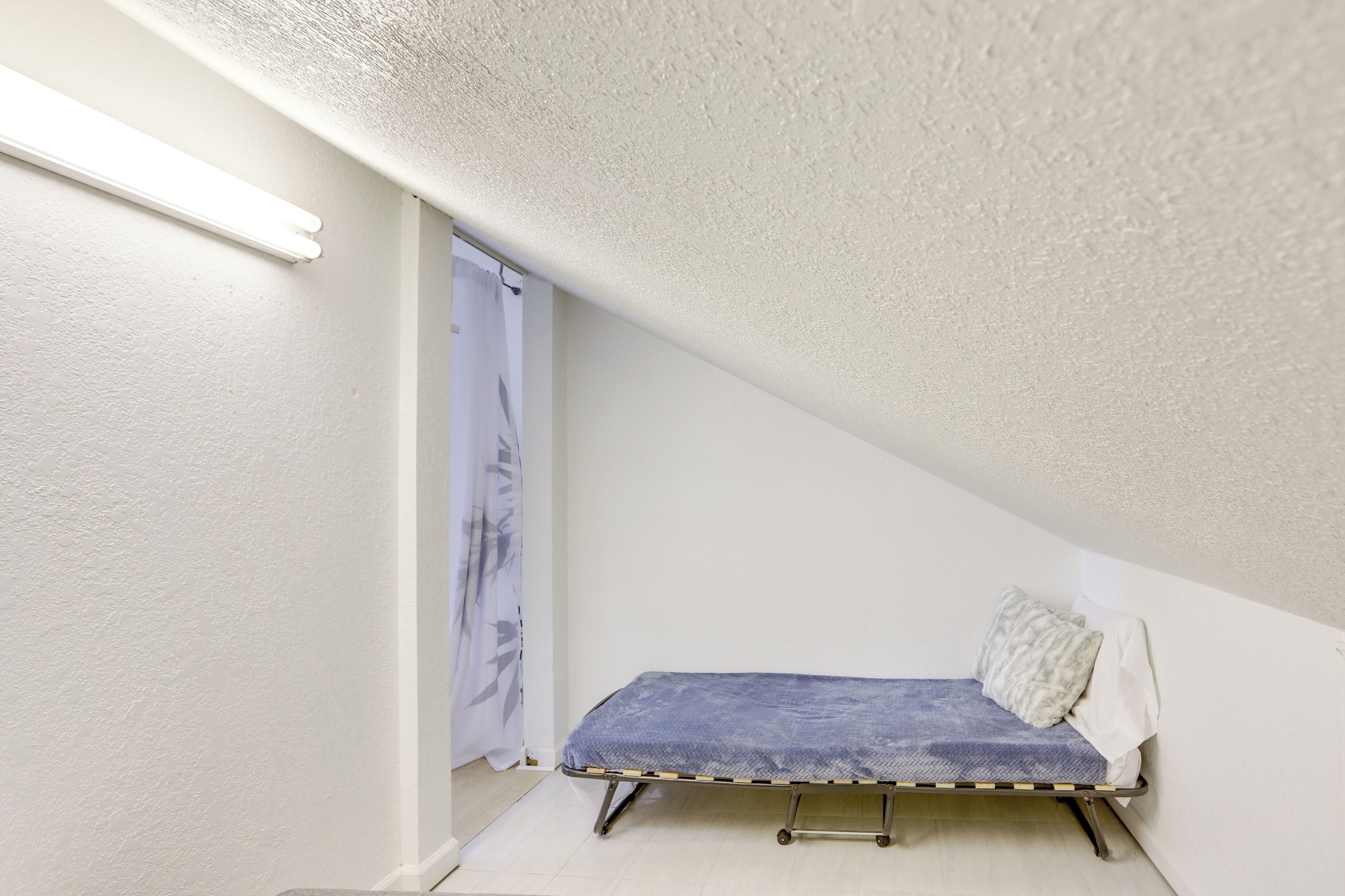A small, single-bed room with a sloped ceiling and a visible door with a curtain.