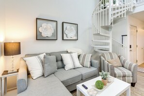 Una sala moderna con un sofá gris, un sillón a rayas y una mesa de centro blanca. Hay dos cuadros florales enmarcados en la pared, una lámpara sobre una mesita auxiliar y una escalera que lleva a un nivel superior.