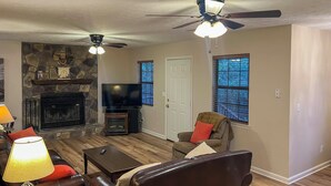 A living room with a stone fireplace, leather sofas, a television, and a ceiling fan. - Creekfront Cabin Near Chattanooga w/ Hot Tub! (Ooltewah)