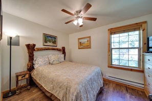A bedroom with a wooden headboard, a bed with a patterned bedspread, a ceiling fan, a window with blinds, and a nightstand with a lamp. - Cozy Countryside Cabin in Robie Creek Park! (Boise)