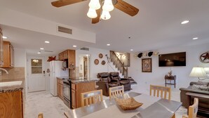 A spacious living area with a dining table, chairs, a sofa, and a TV. There is a kitchen with wooden cabinets and a ceiling fan.