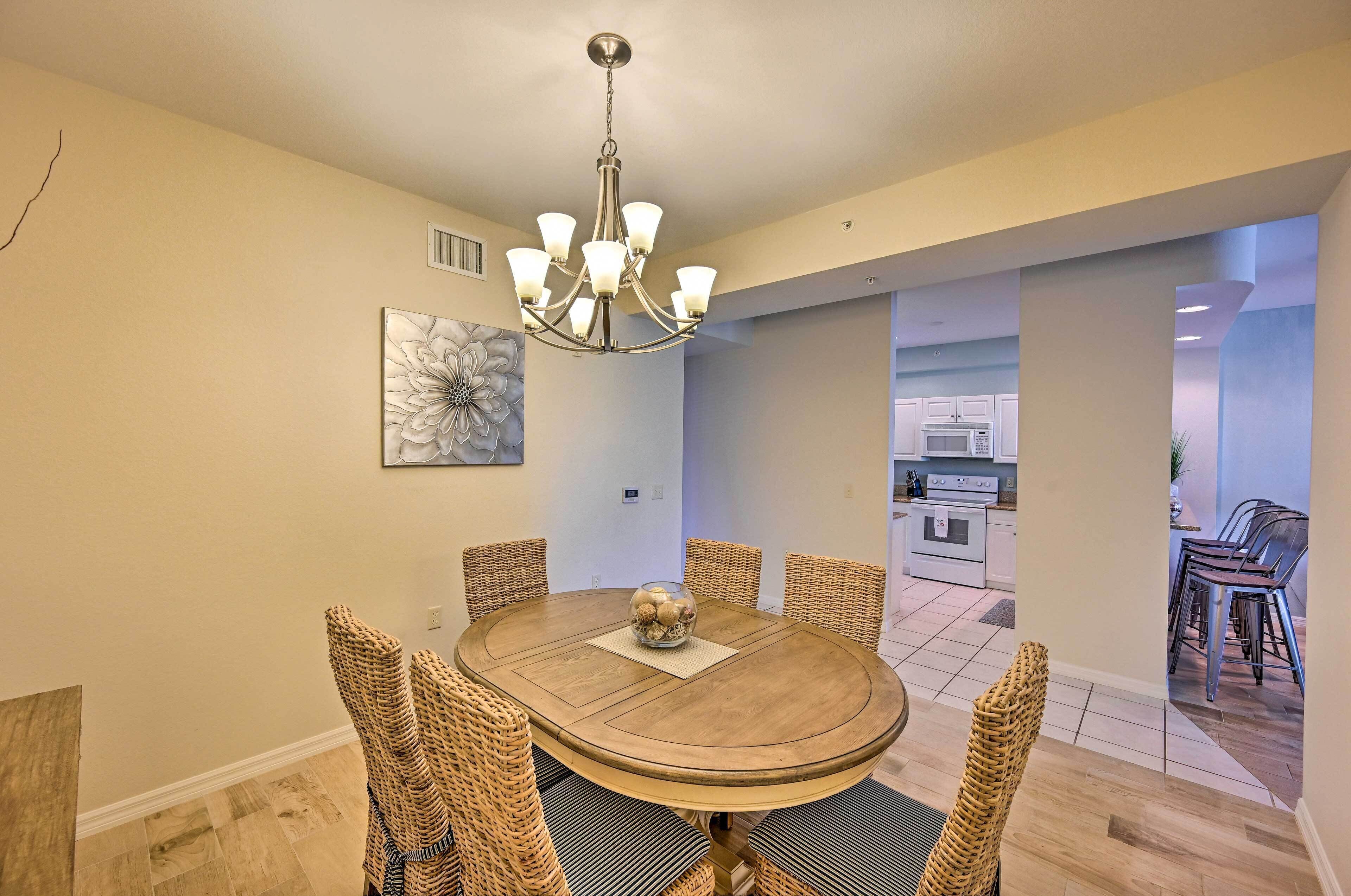 A dining area with a round table, wicker chairs, a chandelier, and a kitchen in the background.