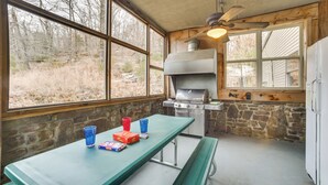 Une cuisine extérieure rustique, avec un mur en pierre, un plafond en bois et une grande fenêtre donnant sur une zone boisée.