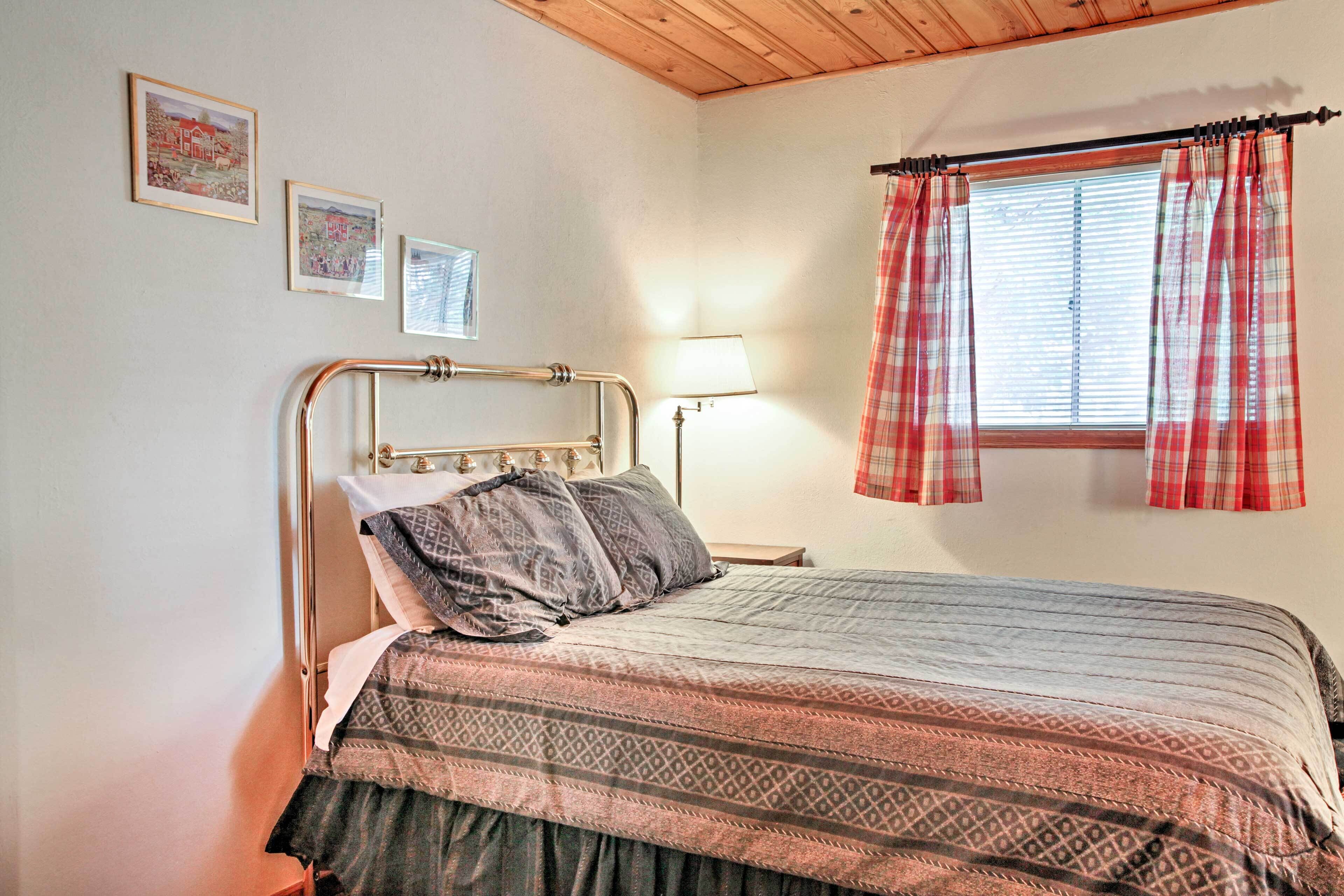Un dormitorio con estructura de cama metálica, ropa de cama estampada, una lámpara de noche y una ventana con cortinas escocesas.
