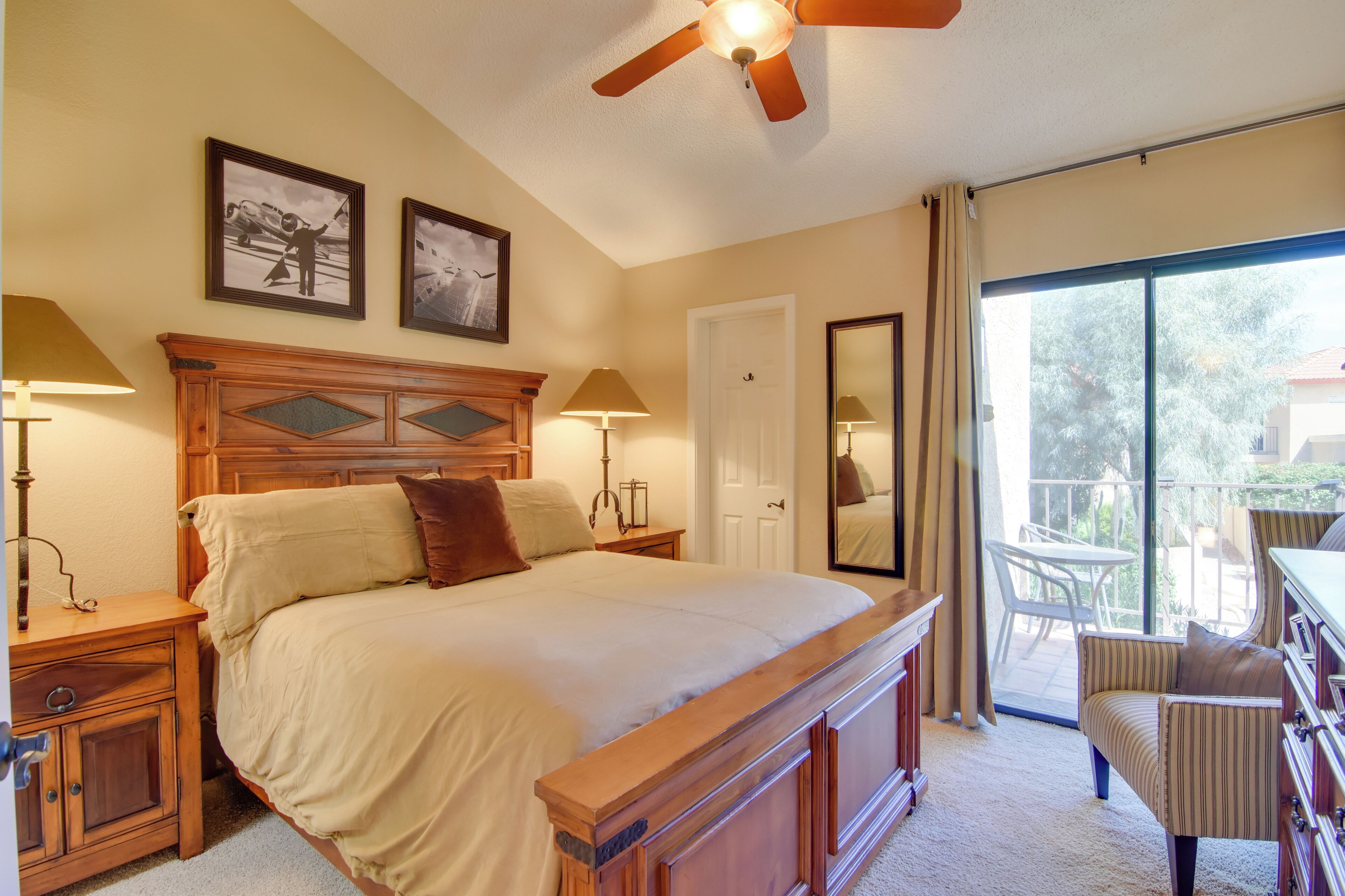 A bedroom with a large bed, wooden headboard, two bedside lamps, a ceiling fan, and a balcony with a chair.