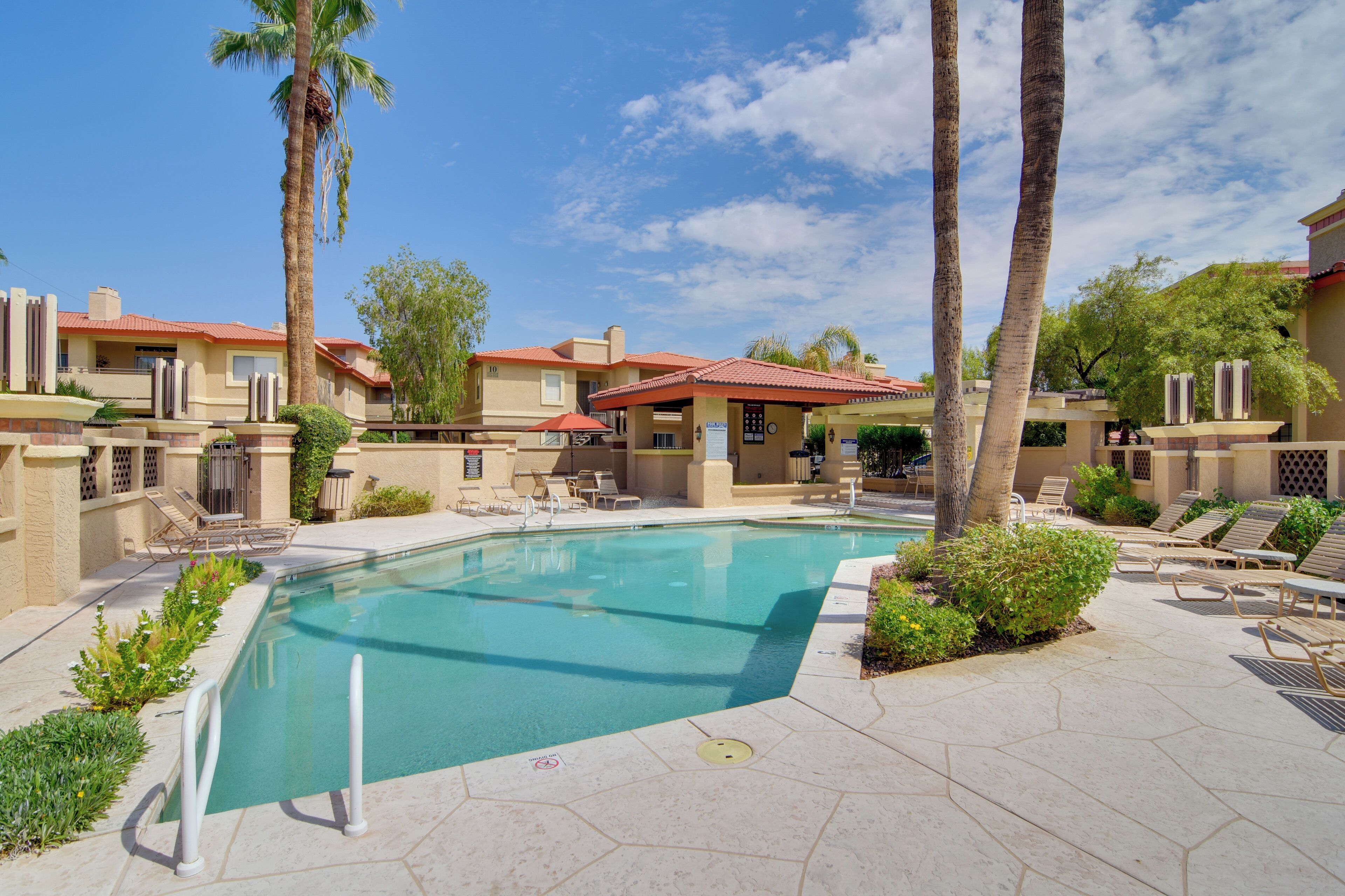 A swimming pool with lounge chairs and palm trees, surrounded by buildings with red roofs.