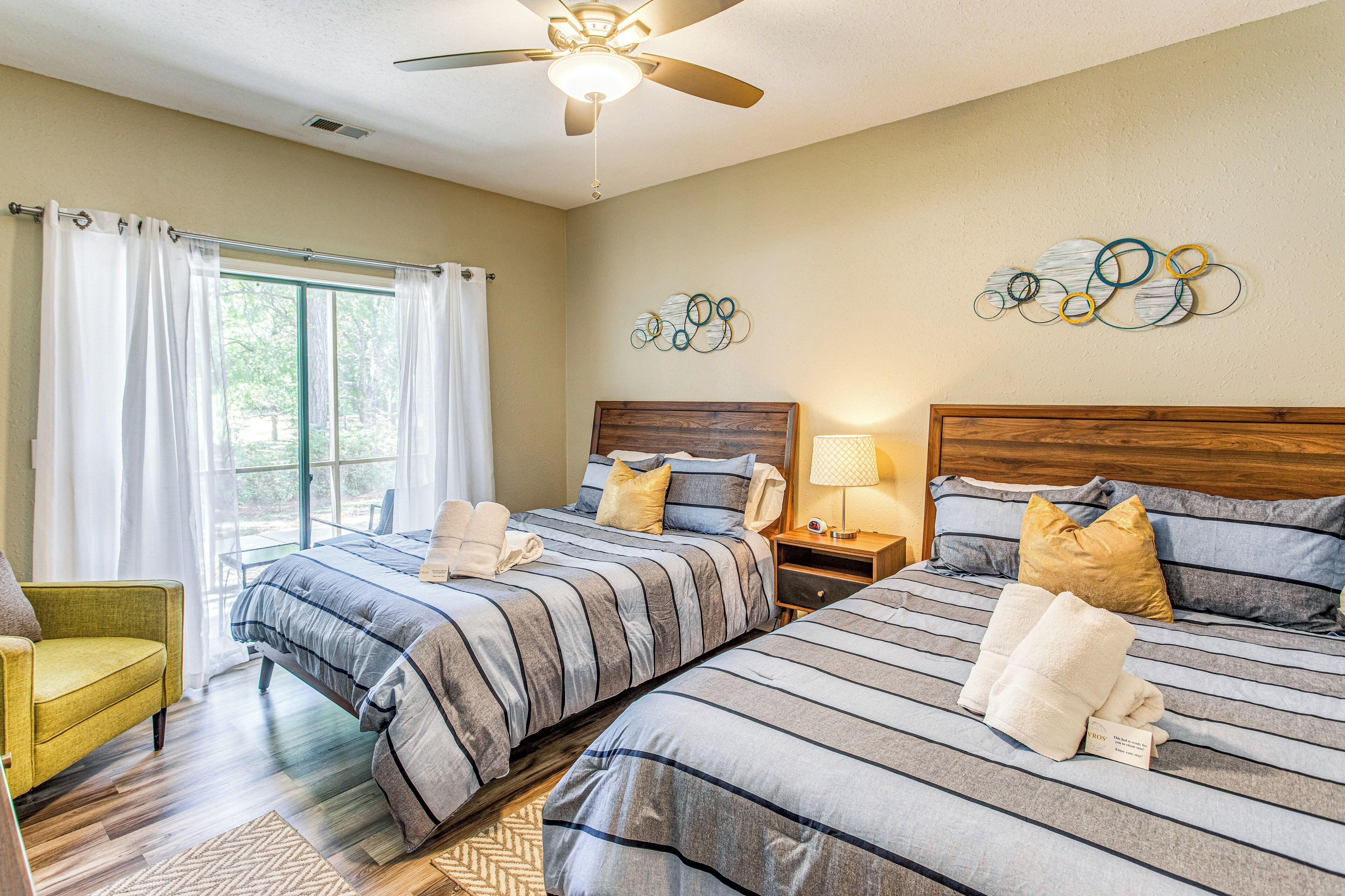 A hotel room with two beds, a ceiling fan, a window with curtains, a wooden headboard, and a wall art piece.
