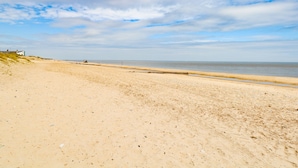 A wide sandy beach with a clear sky and a few clouds. There are a few buildings and a lighthouse in the distance. - Donnys Plaice (Great Yarmouth)