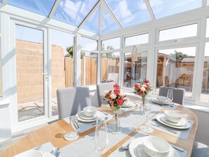 Une véranda avec une table à manger pour quatre personnes, entourée de grandes fenêtres et offrant une vue sur un mur de briques et un jardin.