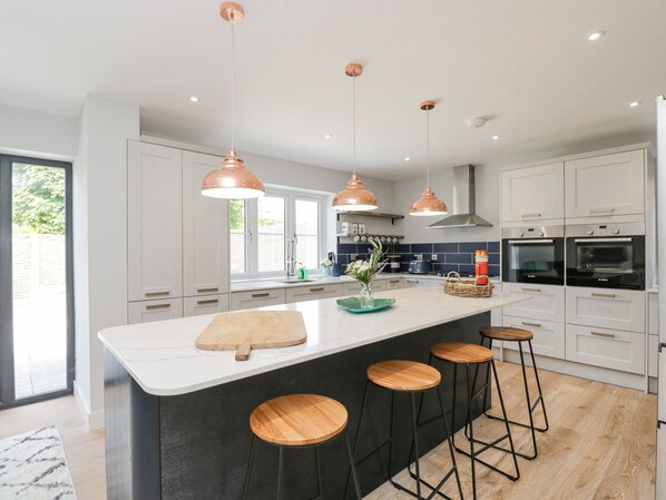 A modern kitchen with white cabinets, a large island with stools, copper pendant lights, and built-in ovens. - Greystones (Gloucester)