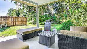 Eine Terrasse im Freien mit Korbmöbeln, einem Grill und einem überdachten Bereich mit Blick auf einen üppigen Garten und einen Holzzaun.