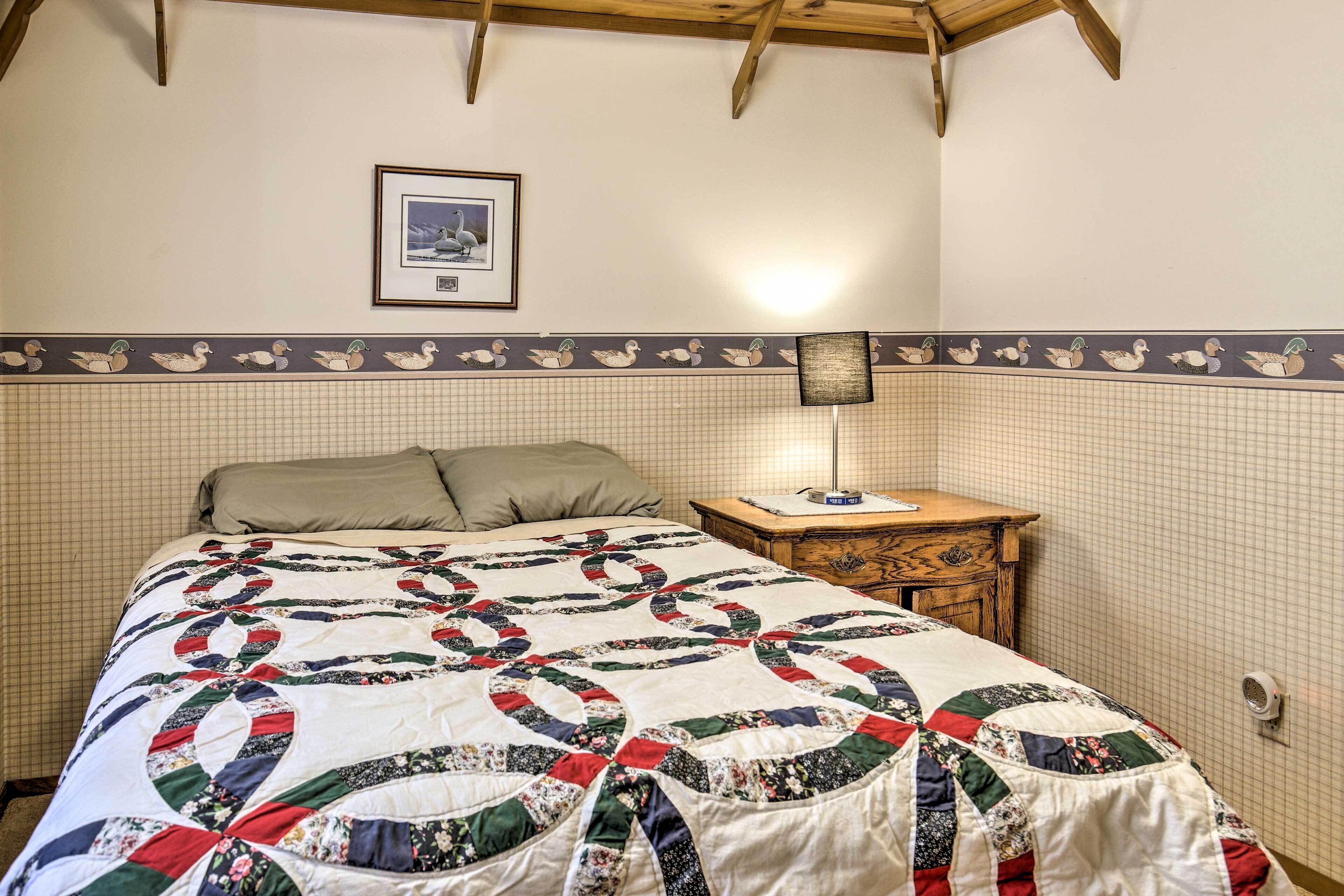 Un dormitorio con colcha acolchada, cabecero de madera, mesita de noche con lámpara y un cuadro enmarcado en la pared.